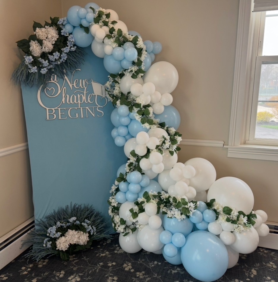 Light blue and white balloon arch with greenery and A New Chapter Begins sign